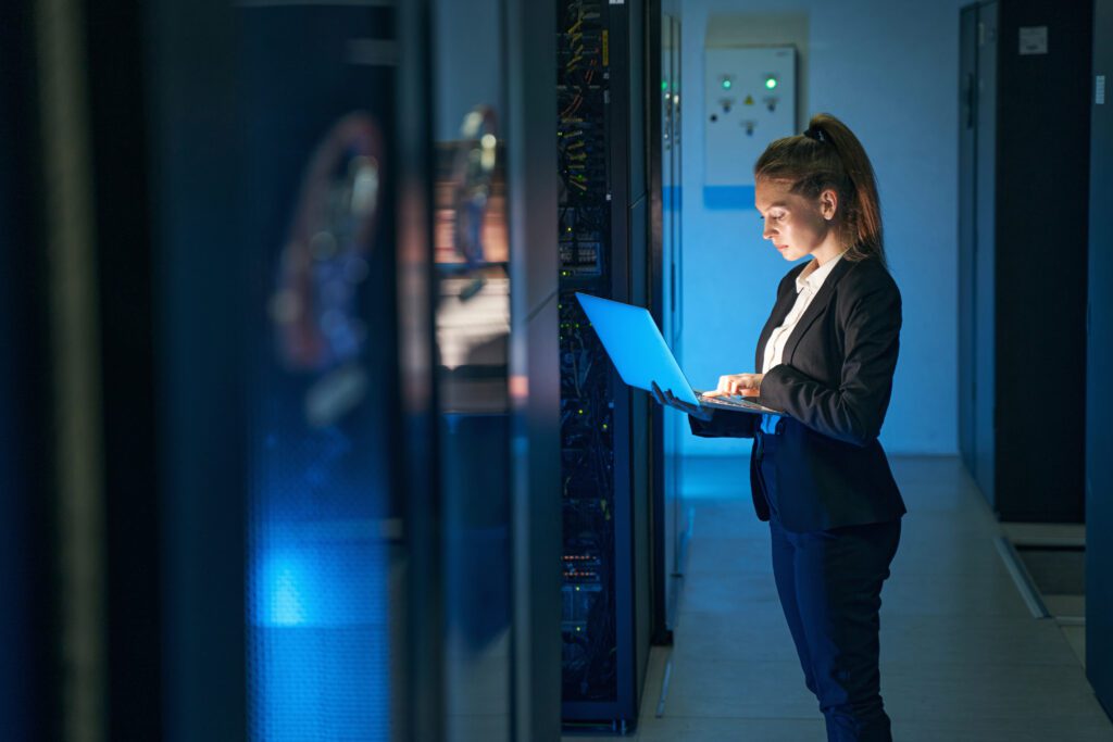 A woman standing in a server room holding a laptop and working on it.