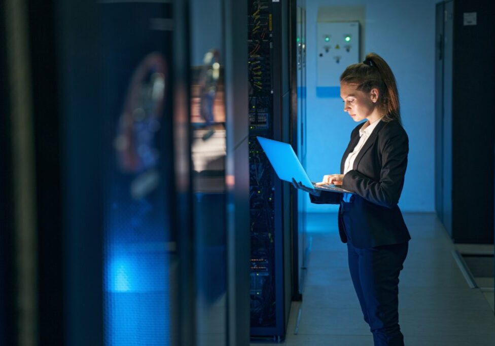 A woman standing in a server room holding a laptop and working on it.